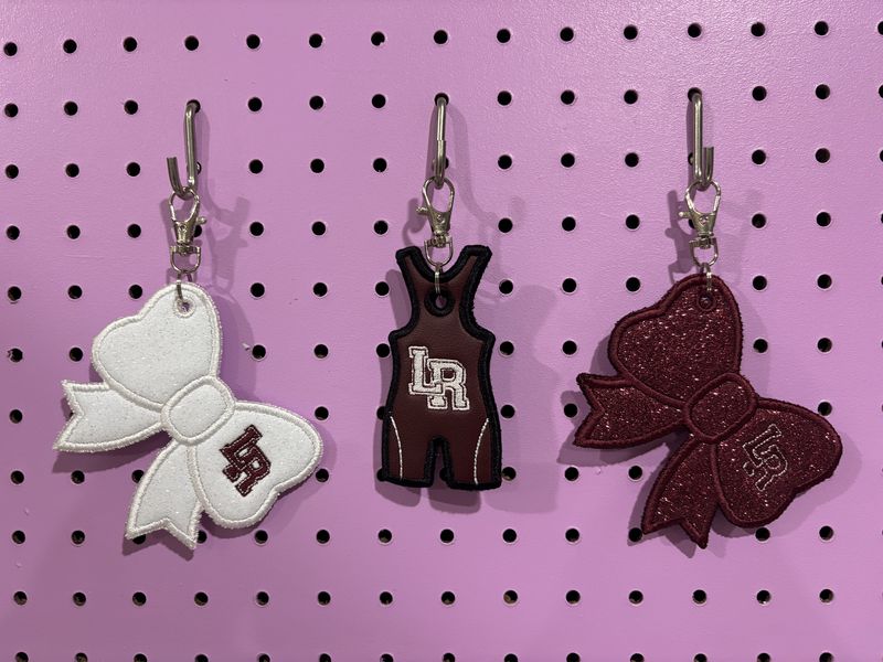 Three LR bag tags on a pegboard: a white glitter bow, a maroon wrestling singlet, and a maroon glitter bow
