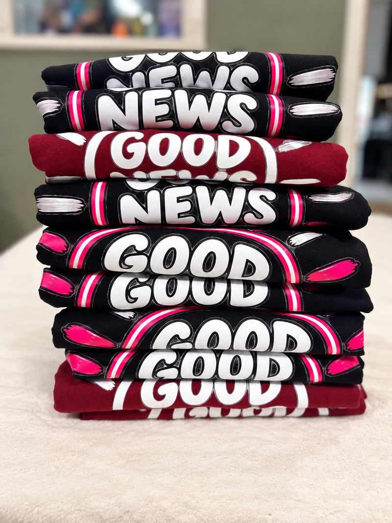 Stack of folded Good News Club shirts in black and maroon with bold white lettering and hot pink accents