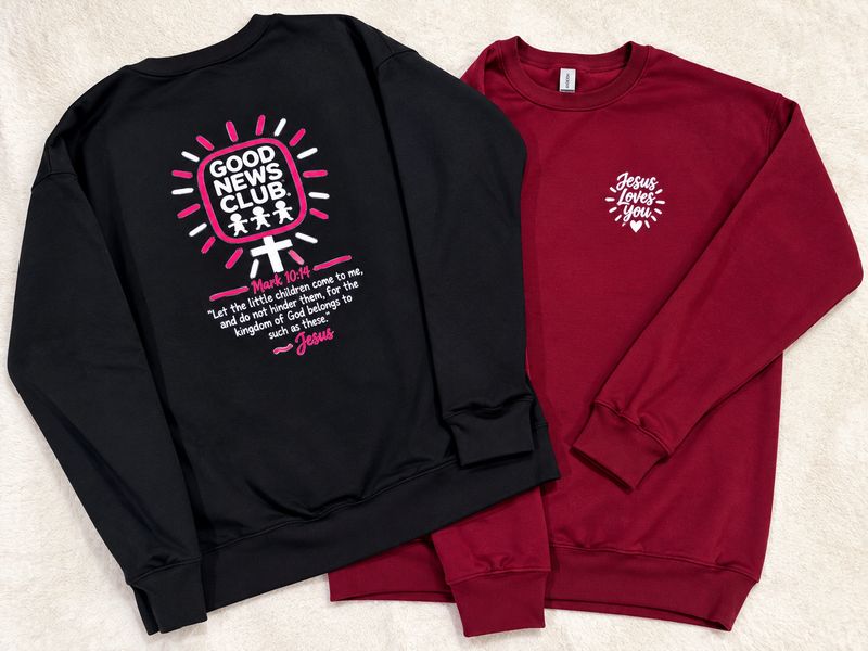 Black Good News Club sweatshirt with a Mark 10:14 verse on the back, beside a maroon sweatshirt with a small Jesus Loves You design