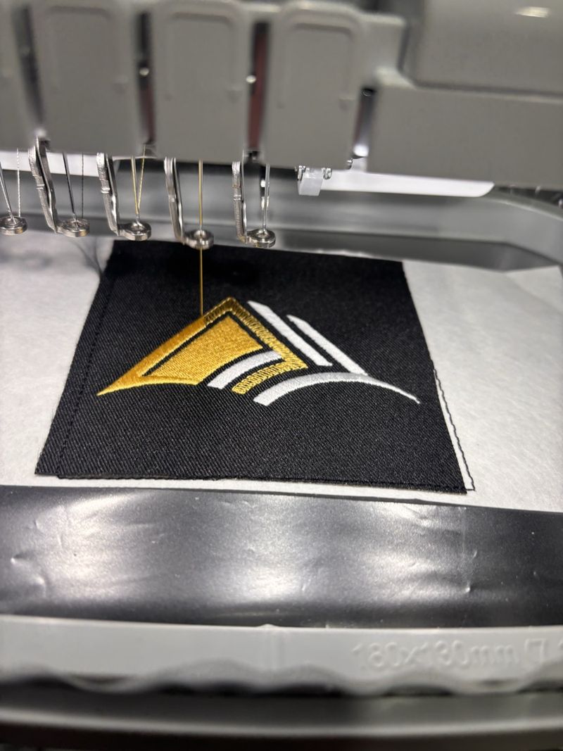 Ten needle embroidery machine stitching a gold and white logo patch