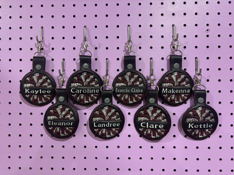 Eight round embroidered cheer name tags in maroon and white, each with a pom pom design and a name, hanging on a purple pegboard