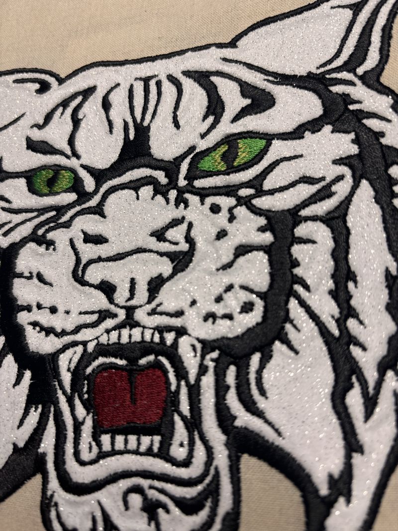 Close-up of the embroidered wildcat showing the sparkly white thread, green eyes, and black outline stitching