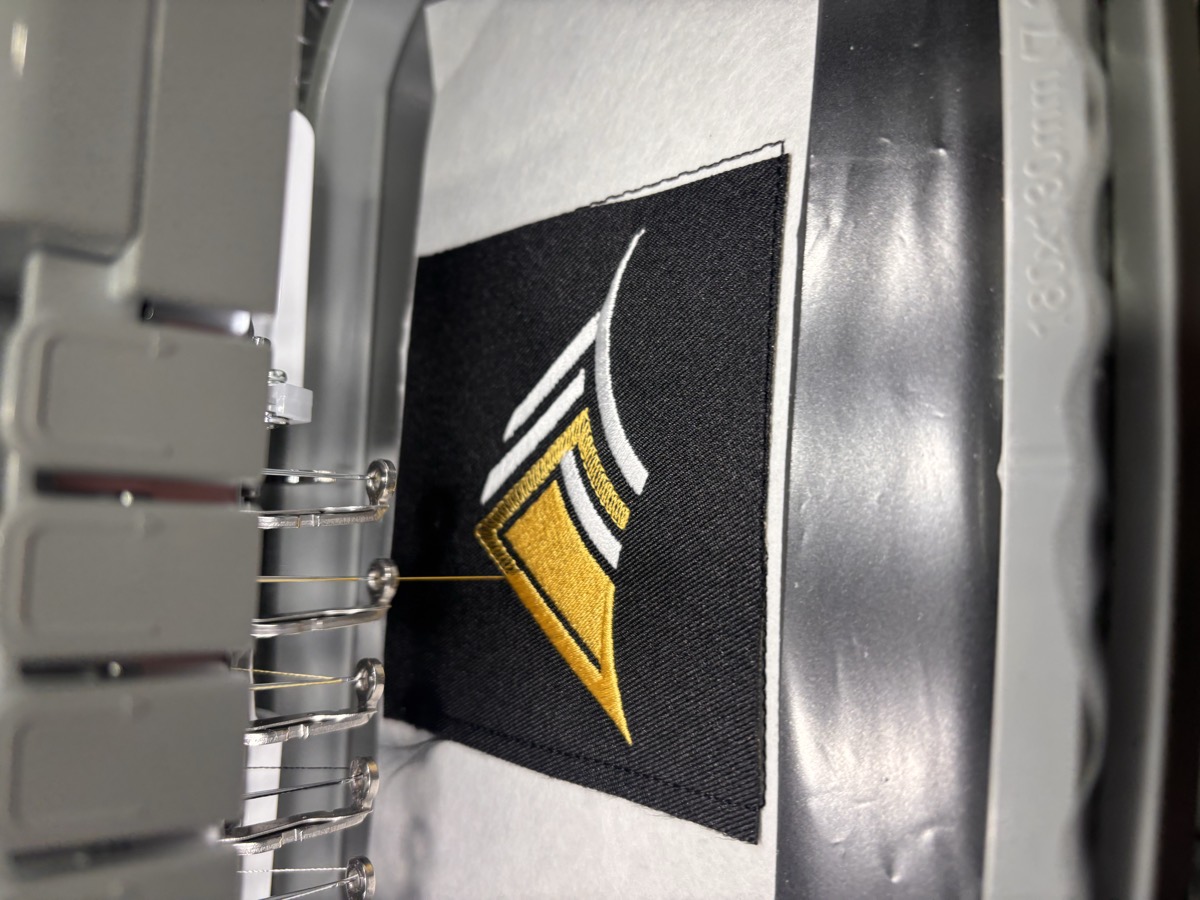Ten needle embroidery machine stitching a gold and white logo patch
